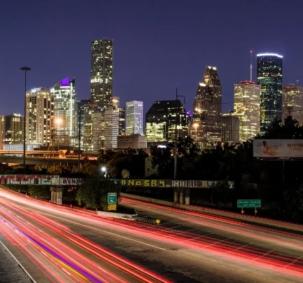 Long exposure photo of city skyline from highway, graffiti on bridges, abstract red lines from car brakes, skyline in background.