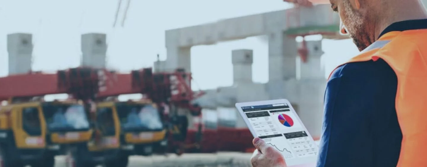 Photo of Construction worker looking at graphs on a computer tablet, construction vehicles in background, worksite in background.