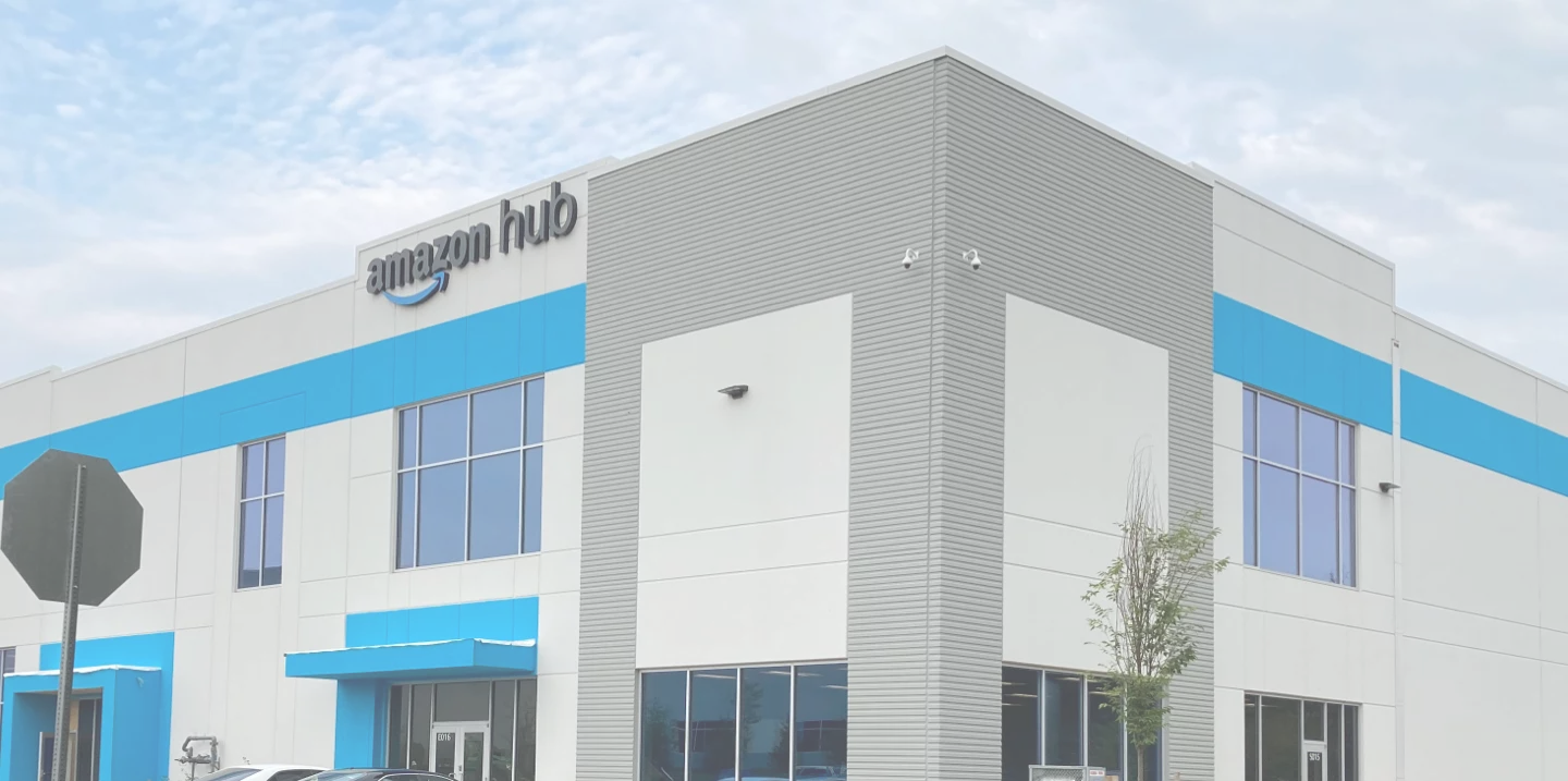 Exterior Amazon Hub Facility, white and gray building with cyan trim, amazon signage at the top of the building, photo taken on a sunny day with clouds.