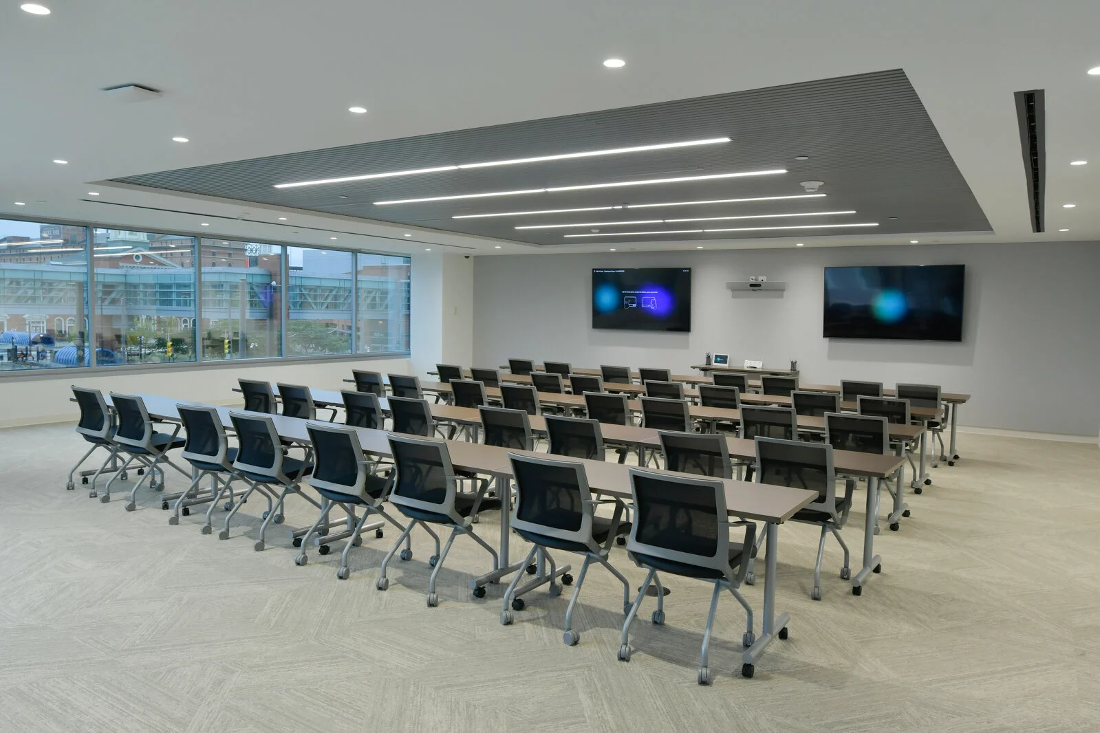 Interior Classroom, 2 tv monitors for presentations, 5 rows of 8 seat desks with computer chairs, carpet floors, large window on left side of the room.
