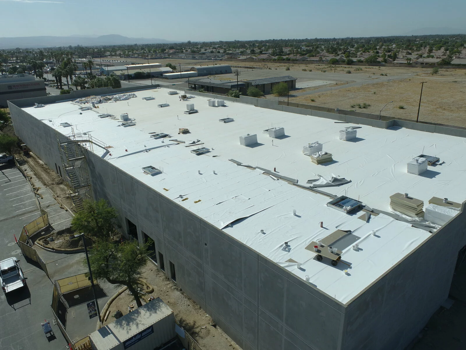 Aerial Exterior of Mathis Brothers Facility, concrete building, building under construction, parking in front, scaffolding, photo taken on a sunny day, trees in the distance.