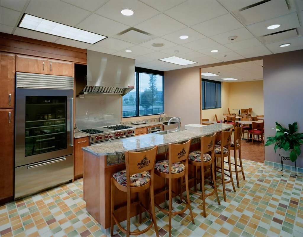 Get Fresh Produce interior community kitchen, multi color tiling flooring, brown cabinetry, granite countertops, large stove, island seating.