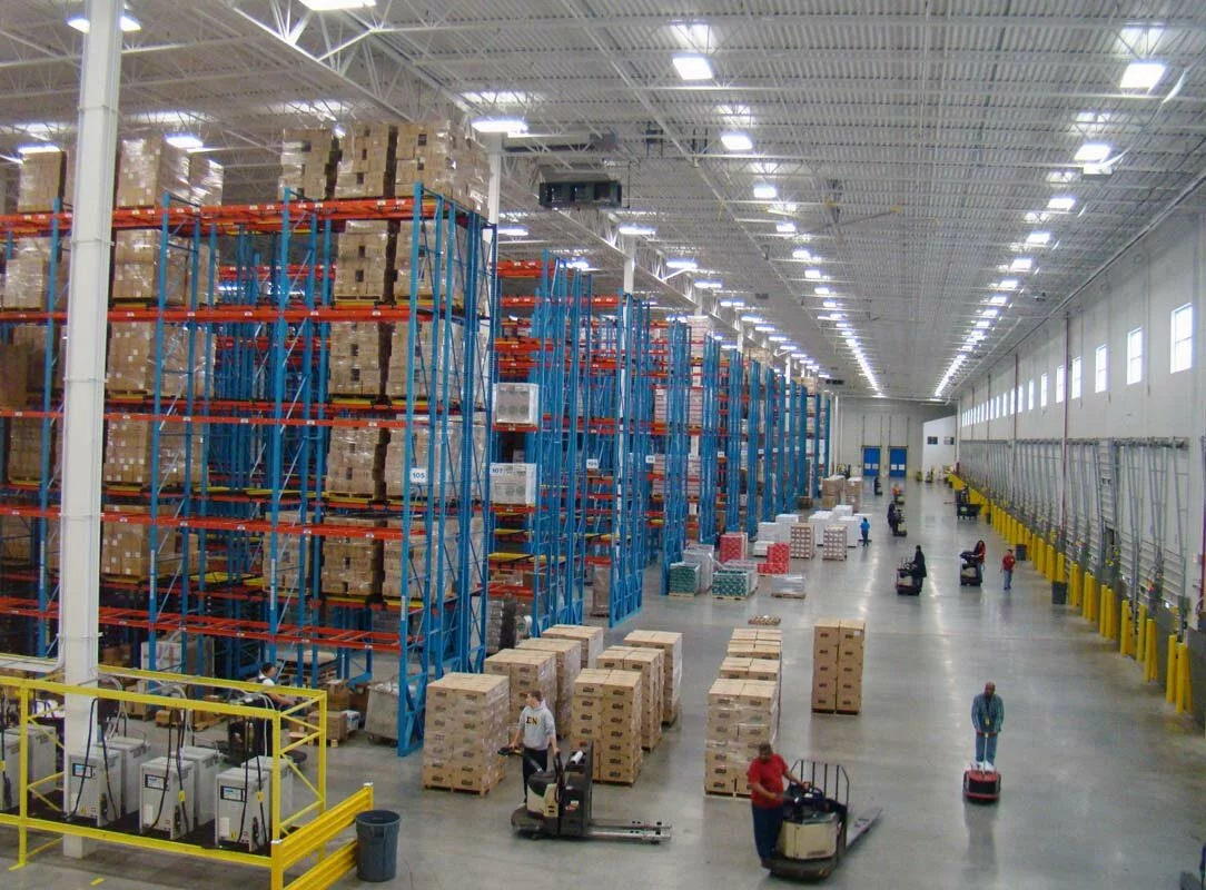 Michael Lewis interior, blue and red shelving, loading bays, product stacks, warehouse machinery and employees working.