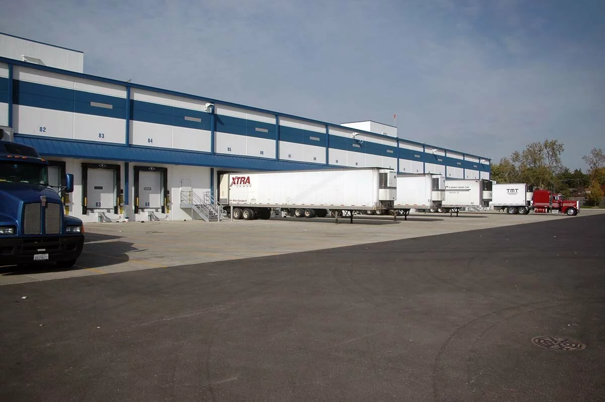 Exterior Sysco Foods, white and blue facility, distribution bays along building, parking lot, photo taken on a cloudy day.