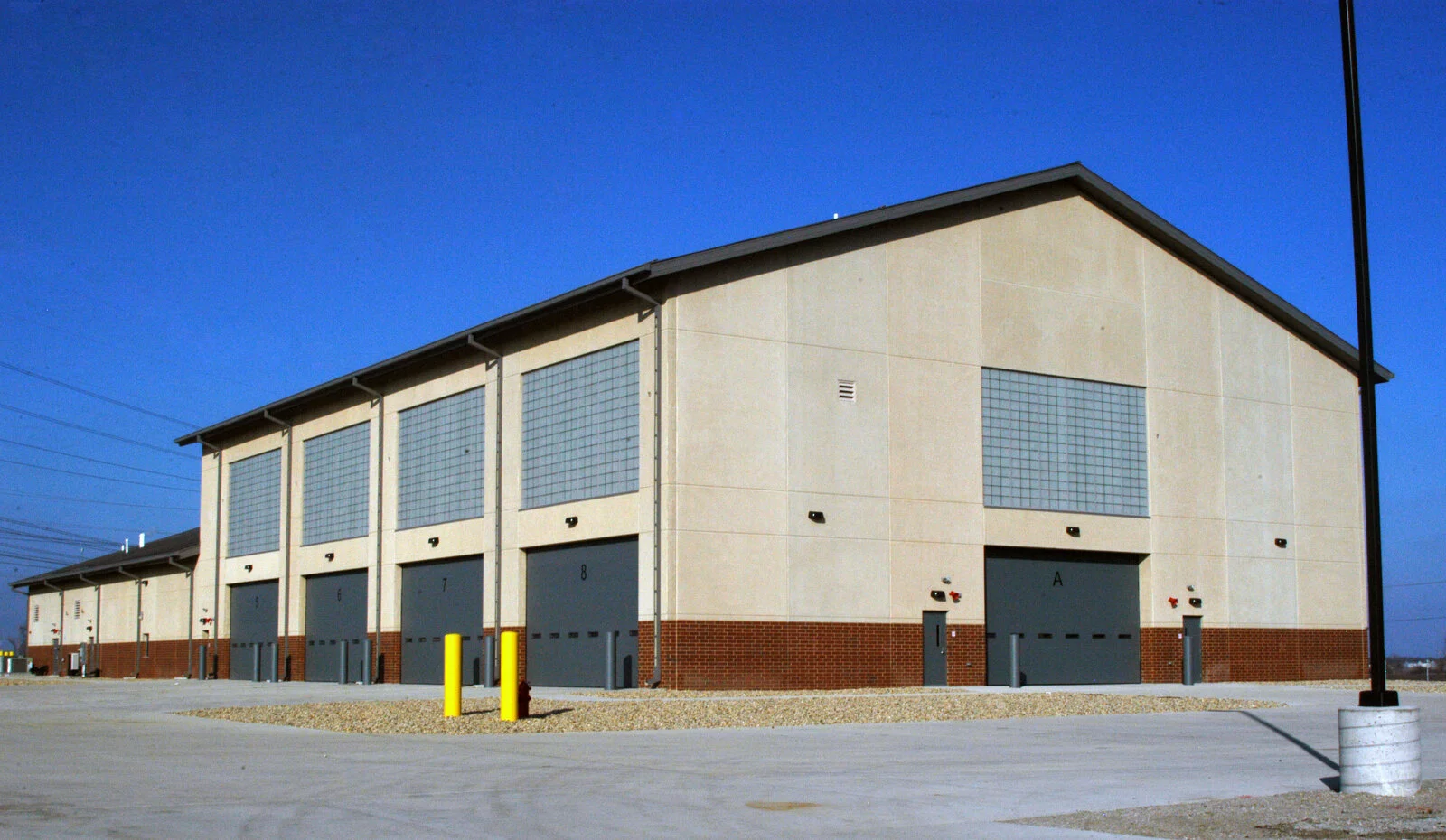 Exterior US Army T.E.M.F. Facility, neutral toned building with red brick at the bottom, large garage doors with equal sized windows above garage doors, bollards around building, photo taken on a sunny day.
