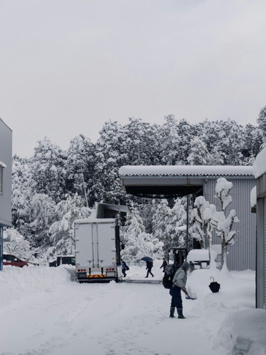 A person is shoveling heavy snow in front of a white delivery truck parked at a loading dock.