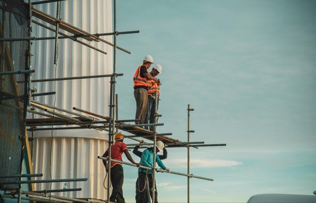 An image showing four construction workers on a multi-level metal scaffolding structure against a clear, light blue sky.