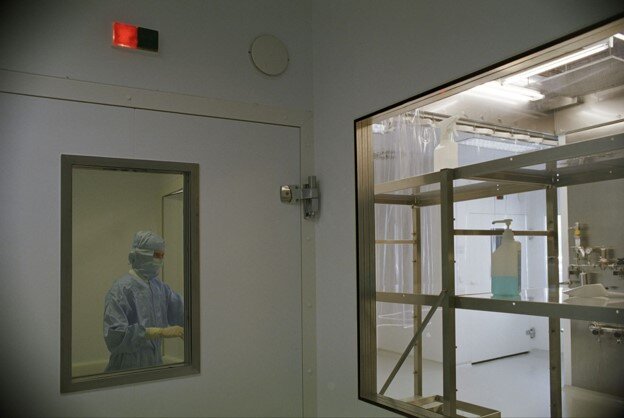 An operator wearing full cleanroom protective garments is visible through an observation window next to an entry pass-through room containing metal shelving units and sanitization supplies.